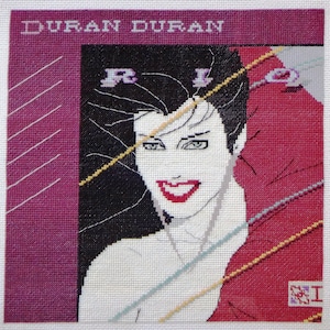 May include: A cross-stitch rendition of the Duran Duran album cover for Rio. The cover features a woman with black hair and a red and white dress. The album title "Rio" is visible in the top right corner.