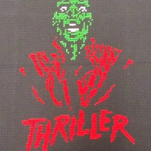 Peut inclure: Broderie au point de croix d'un monstre vert avec une bouche blanche et des mains rouges, avec le mot "THRILLER" brodé en rouge en dessous.