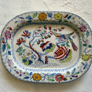 May include: An oval ceramic platter featuring a hand-painted design of a colorful bird perched on a flowering branch. The rim is decorated with a repeating pattern of yellow, pink, and blue flowers and green leaves on a cream-colored base.