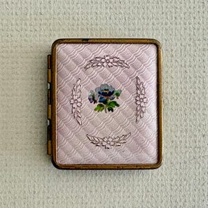 May include: A rectangular metal compact case featuring a pale pink guilloché enamel surface with a wavy, textured pattern. The center displays a small, hand-painted floral motif in blue, green, and yellow, surrounded by four embossed floral sprigs.