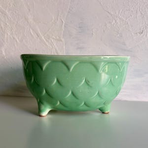 May include: A light mint green ceramic bowl featuring a raised, repeating scalloped pattern across its exterior surface. The bowl is supported by three small, integrated feet and has a smooth, glossy finish throughout its rounded form.
