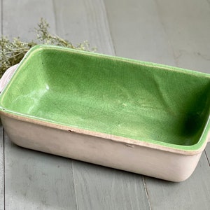 May include: A white ceramic loaf pan with a green glazed interior. The pan has a rectangular shape with rounded corners and two small handles on the sides.