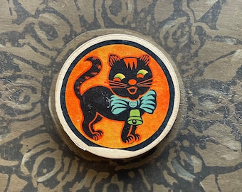 Könnte beinhalten: Ein vintage Halloween-Papieruntersetzer mit einer schwarzen Katze mit einer blauen Schleife und einer Glocke. Die Katze lächelt und hat gelbe Augen. Der Hintergrund ist orange.