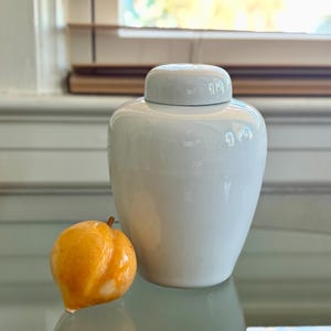 May include: A white ceramic ginger jar with a matching lid, next to a small, orange, fruit-shaped decorative object. The jar has a glossy finish and a classic, rounded shape. The fruit has a smooth texture and a stem.