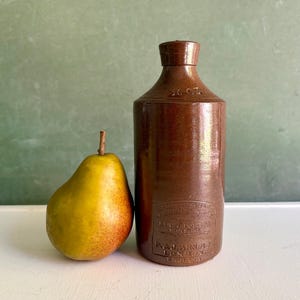 May include: A brown stoneware bottle with a rounded top and a label, next to a yellow and red pear. The bottle has a cylindrical shape and a small opening. The pear is ripe and has a stem. The background is a muted green.