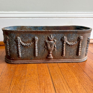 May include: Rectangular, weathered metal planter with a blue-gray patina. The planter features ornate relief detailing, including garland and urn motifs, and sits on a wooden floor. A decorative container for plants.