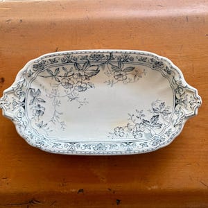 May include: An oval ceramic serving tray with a white base and a dark blue floral pattern. The tray features decorative handles on both ends and a detailed border design with intricate botanical illustrations across the surface.