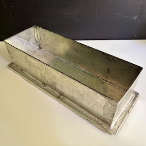 May include: A rectangular metal loaf pan with a weathered, silver-toned finish. The container features straight, slightly angled walls and a flat, extended base rim. The interior surface shows signs of use with visible metallic textures and marks.