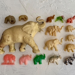 May include: A collection of vintage elephant figurines in various sizes and colors. The elephants range from a large, off-white figurine to smaller, colorful plastic ornaments in shades of pink, green, and brown. Some have detailed carvings, while others are simpler.