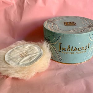 May include: A round, light blue dusting powder container with gold lettering that reads Indiscret DUSTING POWDER and a matching logo. Beside it sits a round, fluffy, cream-colored powder puff with a smooth center featuring the same logo.