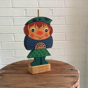 May include: A wooden toy figure of a person with a green hat, blue shirt, and green skirt. The figure is painted with a smiling face and has a flower design on its chest.