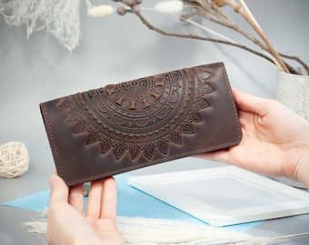 May include: A brown leather wallet with an embossed mandala design. The wallet is rectangular and has a detailed, symmetrical pattern. The leather appears to be a rich, dark brown color. The wallet is held in front of a soft background.