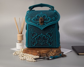 May include: Teal leather handbag with embossed floral design and a top handle. The bag features a brass clasp and is displayed on a wooden base. Decorative items like pearls, a key, and a diffuser are also present.