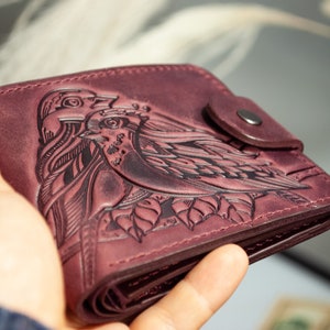 Puede incluir: Cartera de cuero burdeos con un diseño en relieve detallado de dos pájaros y follaje. La cartera tiene un cierre de botón a presión y se sostiene abierta, mostrando el interior. El cuero tiene un aspecto envejecido.