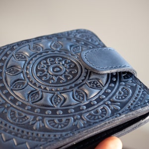 Puede incluir: Cartera de cuero azul con un diseño de mandala floral en relieve. La cartera tiene un cierre de botón a presión y un patrón detallado. El color es un azul profundo y apagado, y el cuero parece texturizado.