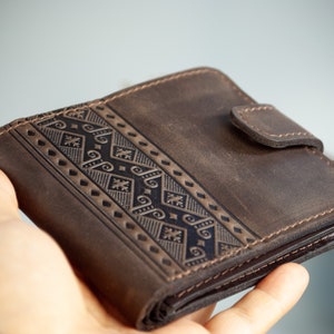 Könnte beinhalten: Eine braune Lederbrieftasche mit einem dekorativen geometrischen Muster. Die Brieftasche hat einen Schnappverschluss und wird in einer Hand gehalten. Die Brieftasche ist etwa 10 cm breit und 10 cm hoch.