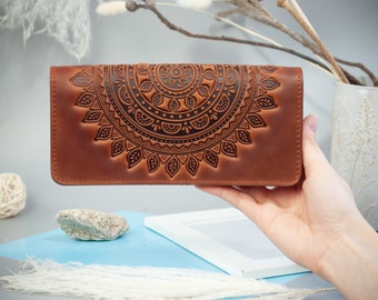May include: A brown leather wallet with an embossed mandala design. The wallet is rectangular and features a detailed, symmetrical pattern across the front. The color is a warm, medium brown, and the design is darker, creating a contrast.