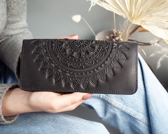 May include: Black leather wallet with an embossed mandala design. The wallet is rectangular and features a detailed, symmetrical pattern across the front. The wallet is held in a person's hands, with a neutral background.