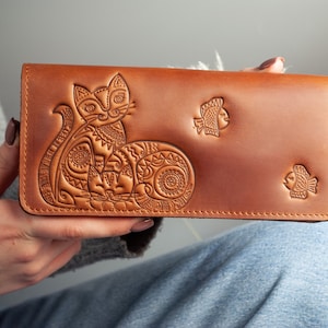 Könnte beinhalten: Eine braune Ledergeldbörse mit einem geprägten Design einer Katze und zwei Fischen. Die rechteckige Geldbörse hat einen Druckknopfverschluss. Das Katzendesign ist detailliert.