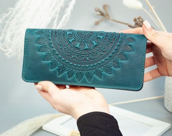 May include: Teal leather wallet with a detailed, embossed mandala design. The wallet is rectangular and features a dark teal color with a contrasting darker embossed pattern. The design is centered on the front of the wallet.