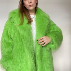 May include: A long, vibrant green faux fur coat. The coat has a wide collar and long sleeves. The texture of the fur is visible. The coat is open, revealing a white top underneath. The person is wearing rings on their fingers.
