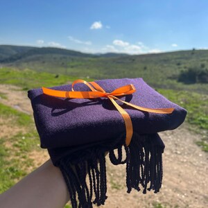 May include: A purple scarf with a bright orange ribbon tied around it. The scarf is folded and has fringe on the edges.