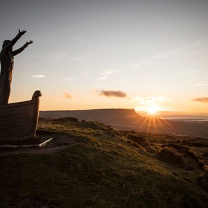 May include: A silhouette of a statue of a person standing in a boat with arms raised, facing a sunset over a mountain range and a body of water.