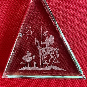 May include: A triangular glass paperweight with an etched design of a person riding a horse. The design is in a minimalist style with a sun and windmills in the background.
