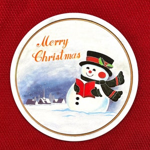 May include: Round Christmas-themed coaster with a snowman illustration. The snowman wears a black top hat, scarf, and holds a red book. The words "Merry Christmas" are written in orange script above the snowman.