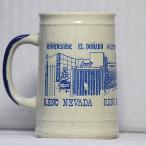 May include: White ceramic mug with a blue handle and blue graphic of the Reno skyline, including the Comstock, Sands, and Harrah's casinos. The text "Riverside El Dorado Hilton Reno Nevada Reno" is printed on the mug.