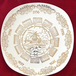 May include: White ceramic plate with gold trim and a calendar design. The plate features a windmill scene and the year 1956.