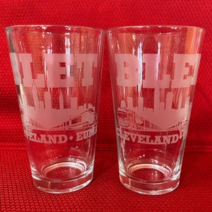 May include: Two clear glass tumblers with etched designs. Each glass features the word "LET" or "BLE" above a skyline and train illustration, with "CLEVELAND" and "EUM" below. The glasses are on a red surface.