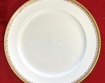 May include: A white porcelain plate with a gold rim and a decorative border of red and gold floral patterns. The plate is round and sits on a red surface.