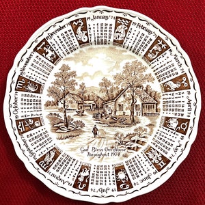 May include: A brown and white ceramic plate with a calendar design featuring the zodiac signs and a rural scene. The plate has a scalloped edge and the text "God Bless Our House Throughout 1974".