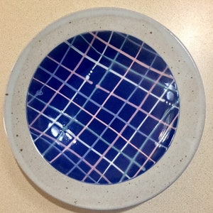 May include: A ceramic plate with a speckled, light gray rim and a deep blue center. The center features a grid pattern of intersecting lines in white, light blue, and pink. The plate is round and appears handmade.