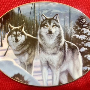 May include: Oval decorative plate featuring two wolves in a snowy forest scene. The wolves have gray and brown fur, with a winter landscape background. The plate has a gold rim and is set against a red background.