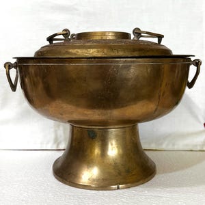 May include: A vintage brass cooking pot with a lid. The pot has a rounded bowl shape, supported by a pedestal base. It features two handles on the sides and a handle on the lid. The brass has a warm, aged patina.