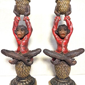May include: Pair of decorative monkey figurines. Each monkey wears a red jacket and holds a basket above its head. The monkeys sit atop a pineapple-shaped base with a gold and brown finish. The figurines are detailed and whimsical.