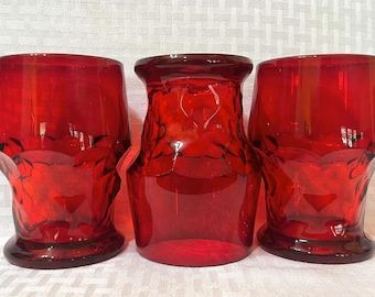 May include: Three vibrant red glass tumblers featuring a distinct textured, faceted surface design. The two outer glasses have a flared rim and a wider base, while the centre glass displays a slightly different silhouette with a smooth, rounded top.