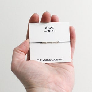 Peut inclure: Un bracelet à cordon noir avec des perles argentées épelant "HOPE" en code Morse est présenté sur une carte blanche. La carte porte également le mot "HOPE" au-dessus du code Morse et le texte "THE MORSE CODE GIRL" en dessous.