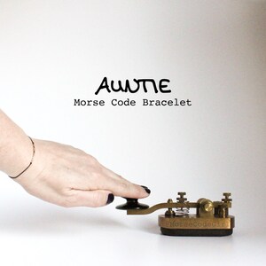Peut inclure: Un bracelet en code Morse épelant "Auntie" est présenté. Le bracelet est porté au poignet, et une main touche une clé télégraphique vintage. Le texte "Auntie Morse Code Bracelet" est au-dessus de la clé.
