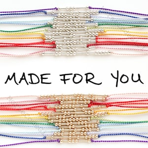 May include: Two rows of colourful string bracelets with silver and gold beads. The bracelets are arranged in a rainbow pattern. The text "MADE FOR YOU" is written in black ink below the bracelets.