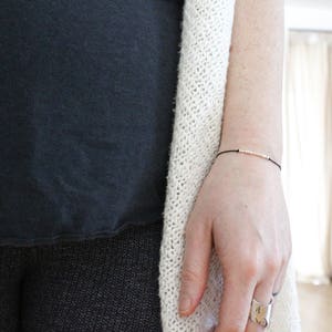 May include: A delicate black string bracelet with small silver and rose gold beads. The bracelet is worn on a wrist, with a silver ring on the finger. The person is wearing a black top and a cream-coloured knitted cardigan.