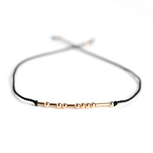 May include: A delicate gold bracelet with small beads and a bar charm on a black cord.