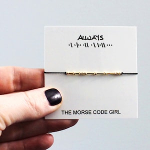 Peut inclure: Un bracelet avec des perles dorées sur un cordon noir, présenté sur une carte blanche. La carte porte le mot "ALWAYS" et des symboles en code Morse. Le texte "THE MORSE CODE GIRL" est imprimé en bas.