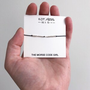 May include: A black string bracelet with silver beads and a silver bar charm. The bracelet is displayed on a white card with the text "HOT MESS... III I II..." and "THE MORSE CODE GIRL".