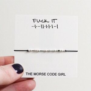May include: A black string bracelet with silver beads spelling out "F**K IT" in Morse code. The bracelet is displayed on a white card with the text "F**K IT" and the Morse code translation. The card also includes the text "THE MORSE CODE GIRL".
