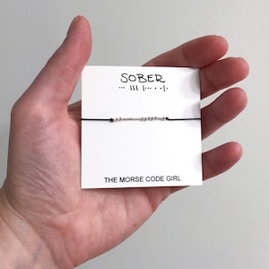 May include: A black string bracelet with silver beads spelling out the word "SOBER" in Morse code. The bracelet is on a white card with the text "SOBER ... ---  . . . - . -" and "THE MORSE CODE GIRL".