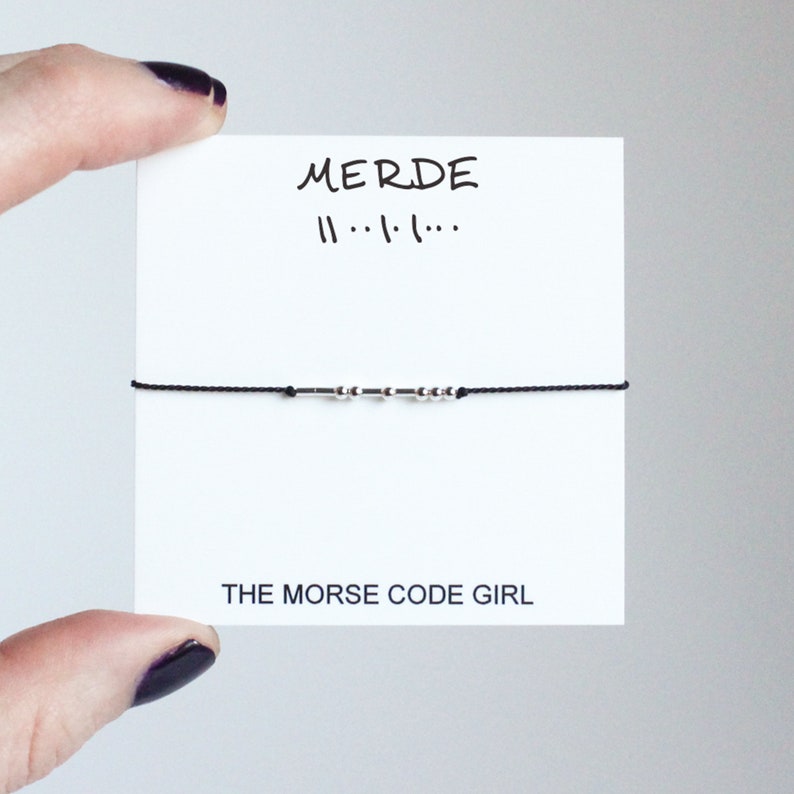May include: A black string bracelet with silver beads spelling "MERDE" in Morse code. The bracelet is displayed on a white card with the text "MERDE" and "THE MORSE CODE GIRL".