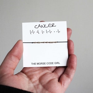 May include: A black string bracelet with gold-coloured beads, presented on a white card. The card displays the word "CANCER" and Morse code symbols. The text "THE MORSE CODE GIRL" is printed at the bottom of the card.